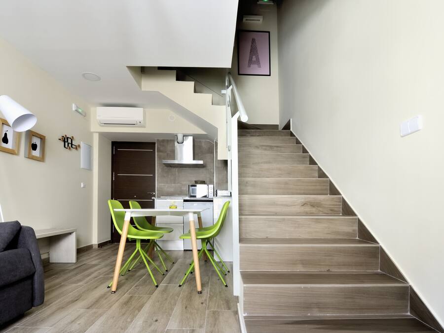 Un espacio habitable moderno con una escalera, un área de comedor con sillas verdes, un sofá gris y una mesa blanca.