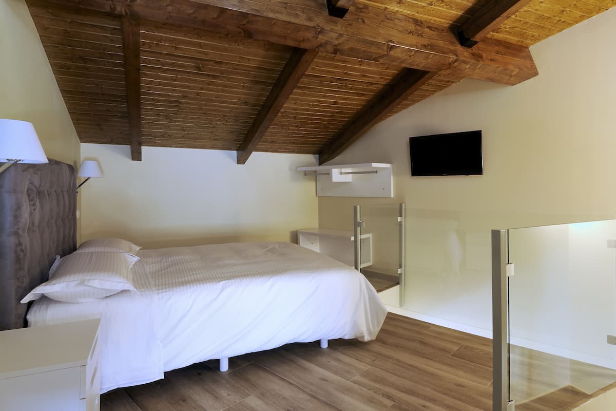 A bedroom with a wooden ceiling, a bed with white bedding, a bedside table, a lamp, a TV mounted on the wall, and a glass partition.