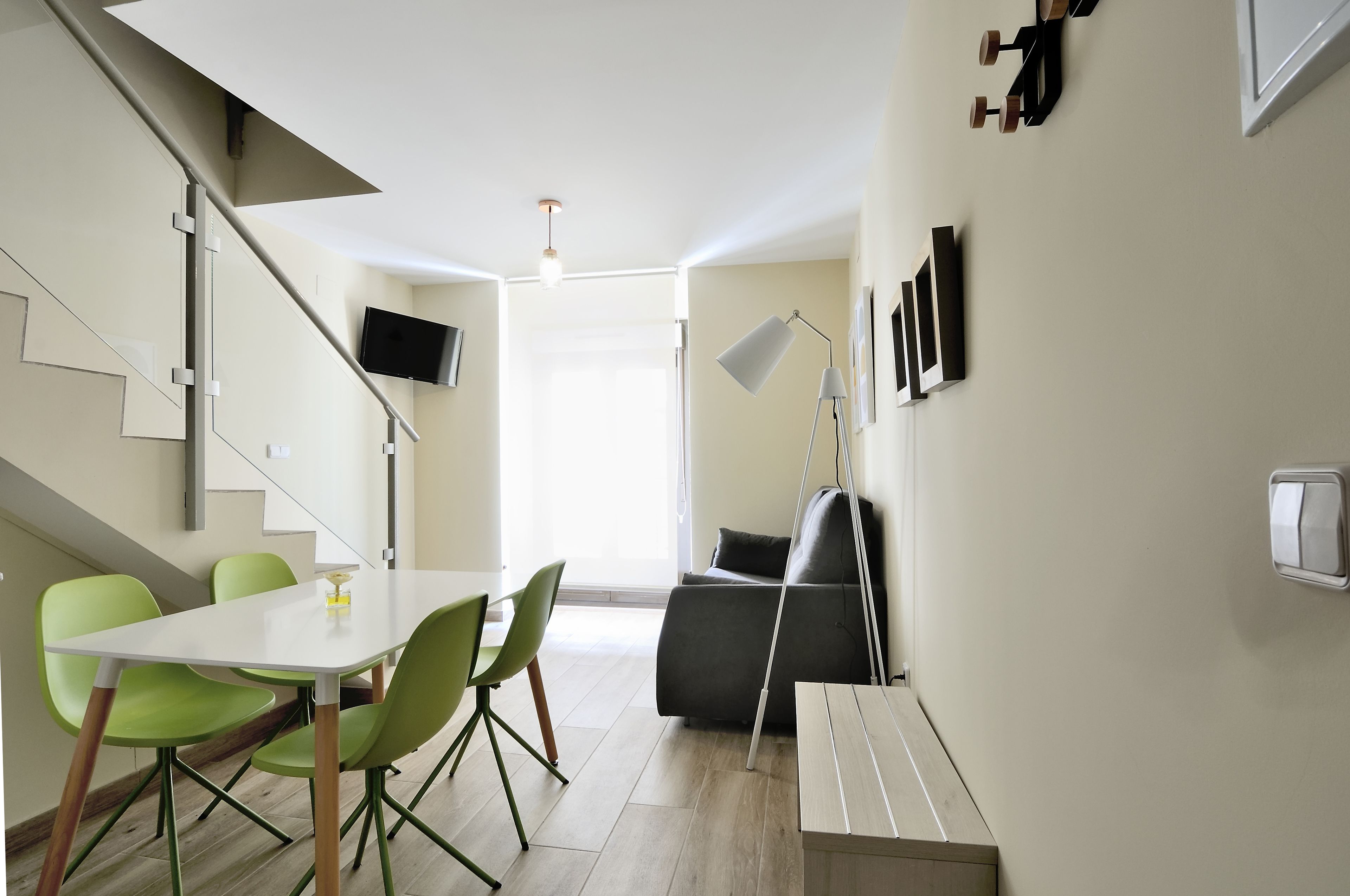A modern living room with a staircase, a dining area with green chairs, a grey sofa, and a flat-screen TV mounted on the wall.