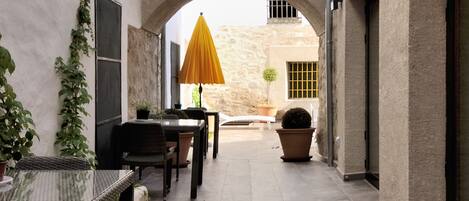 Un patio estrecho con zona de estar techada, una pequeña mesa y plantas en macetas.