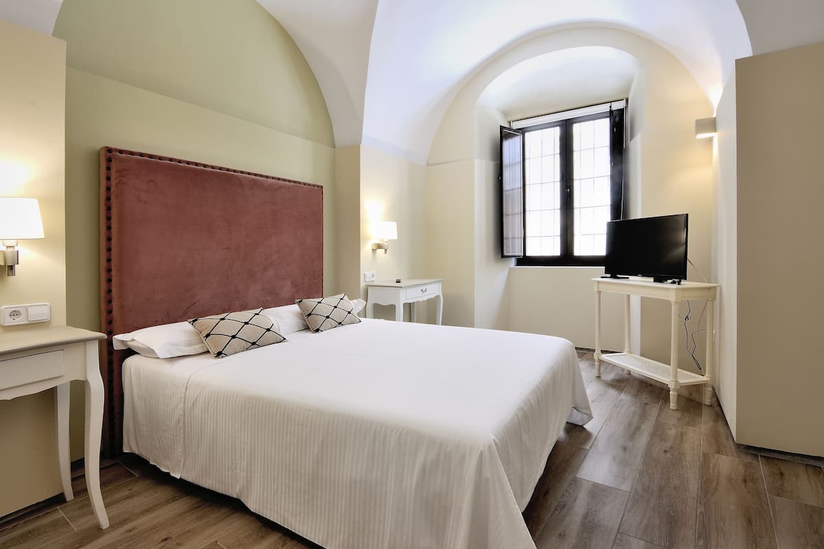 A neatly arranged bedroom with a large bed, a television, and a window providing natural light.