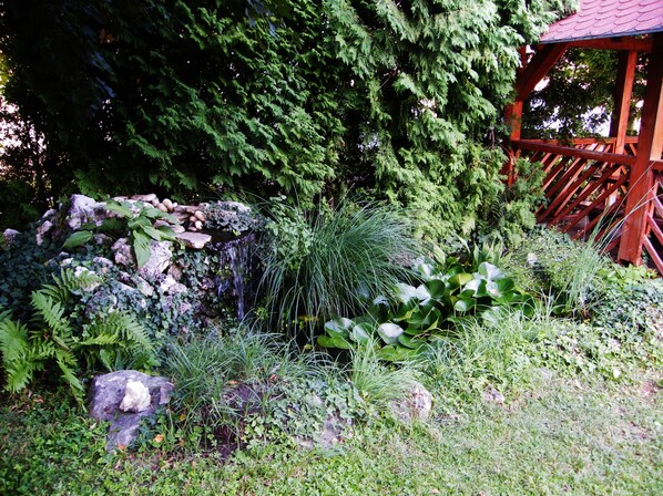 Un jardín con un pabellón de madera, una cascada pequeña y diversas plantas.