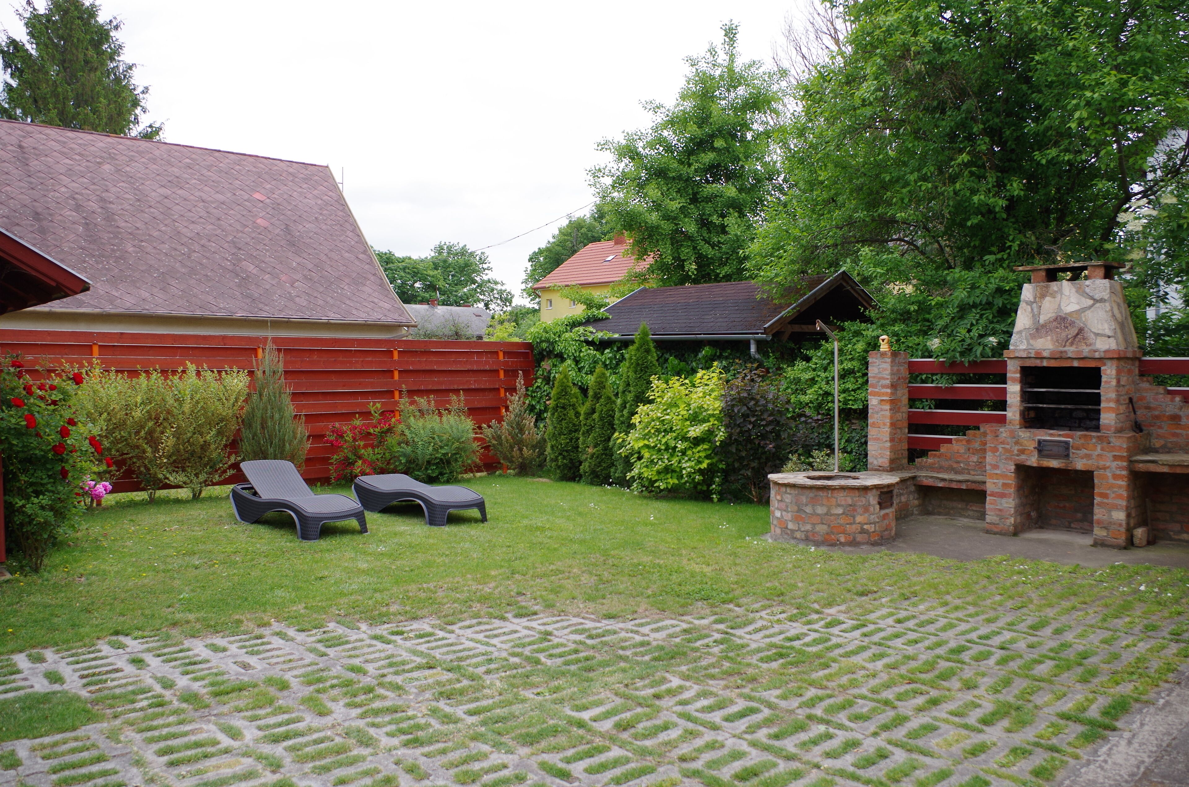 A backyard with a brick barbecue, two lounge chairs, a red fence, and a lawn.