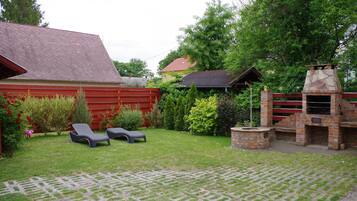 A backyard with a brick barbecue, two lounge chairs, a red fence, and a lawn.