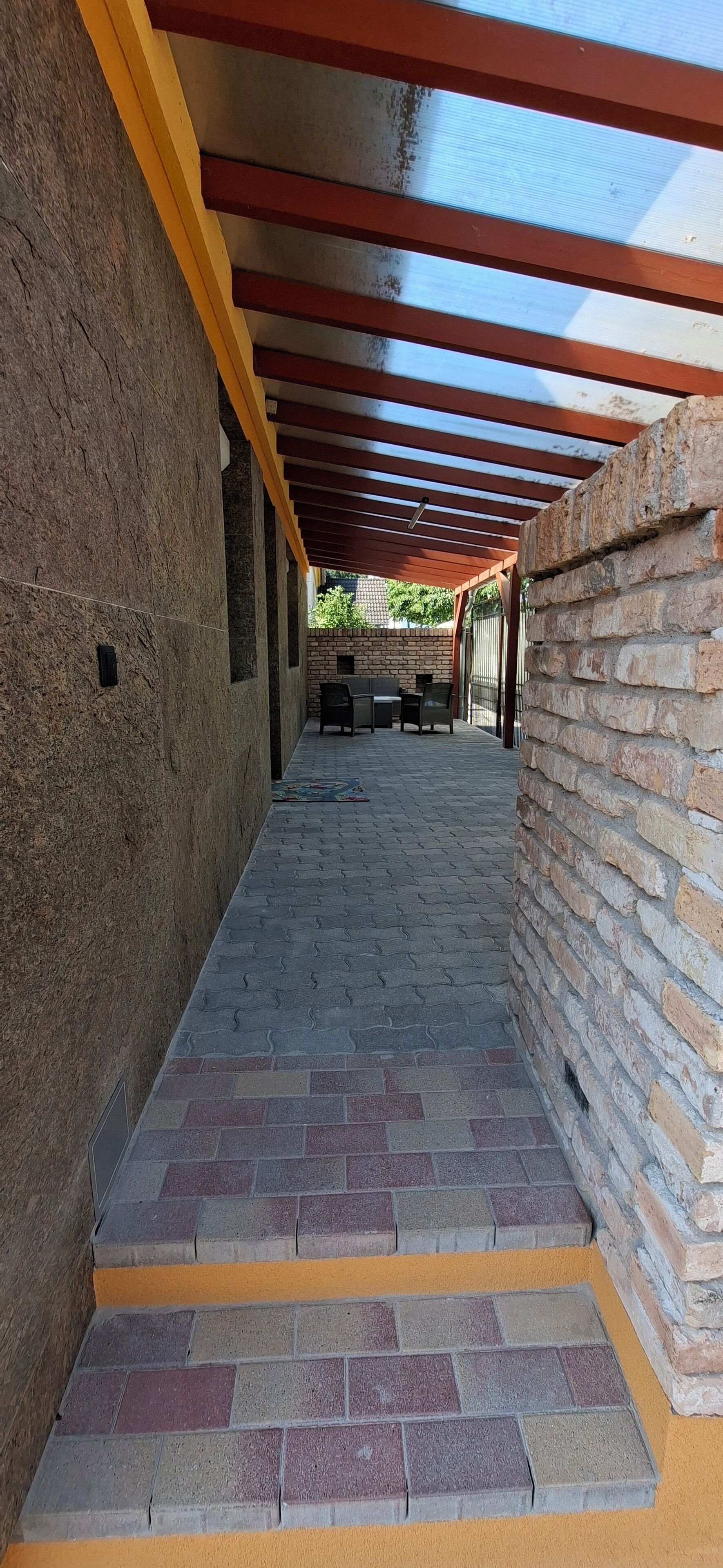 A covered outdoor area with a brick floor, yellow and red beams, and a brick wall.