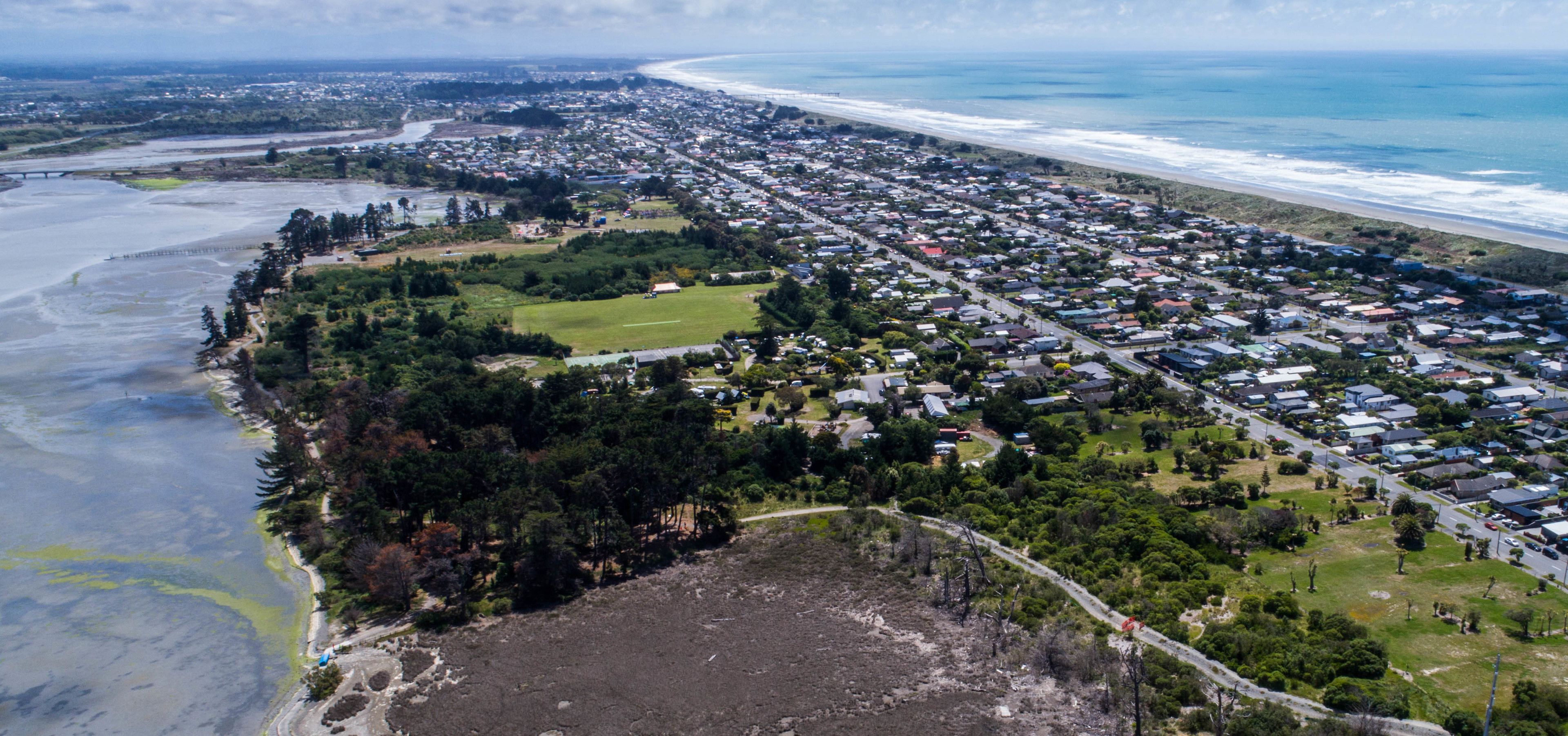 Vista aérea de un pueblo costero con playa, zona residencial y un extenso campo abierto.