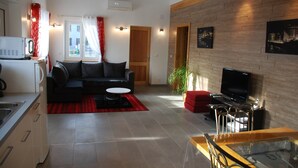 A modern living room with a black sofa, a red ottoman, a glass coffee table, a TV, and a kitchen area with a microwave. - New Kertzfeld cottage, luxury performance in Alsace countryside. (Kertzfeld)