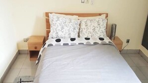 Ein ordentlich bezogenes Bett mit weißer Bettdecke und gemusterten Kissen, ein Nachttisch aus Holz mit einer Lampe und ein kleiner Beistelltisch.