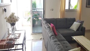 Ein Wohnzimmer mit grauem Sofa, einem kleinen Tisch mit Blumenvase und einer Schiebetür zum Balkon.