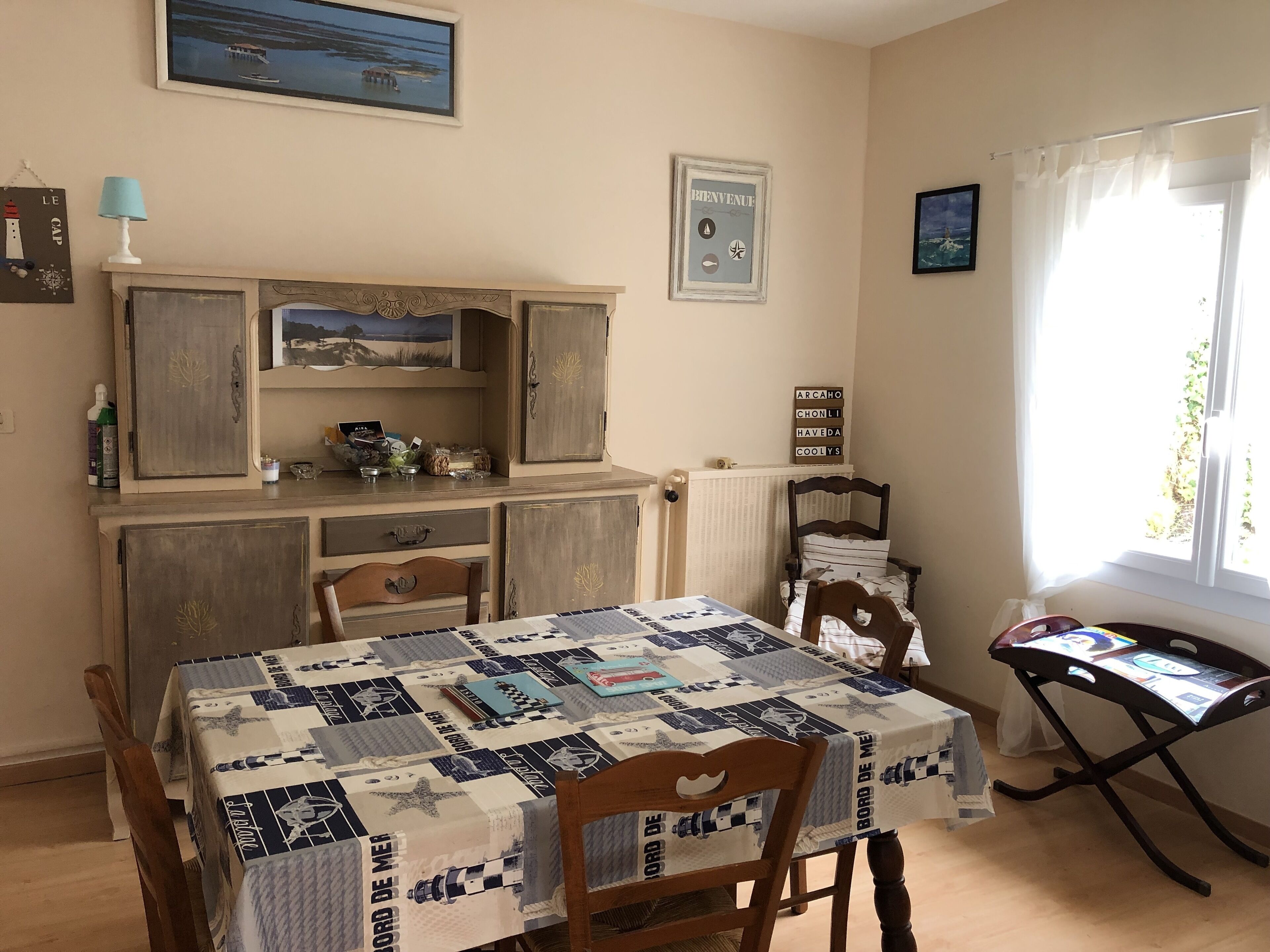 A dining area with a table covered in a nautical-themed tablecloth, wooden chairs, a cabinet with decorative items, and a window with curtains.