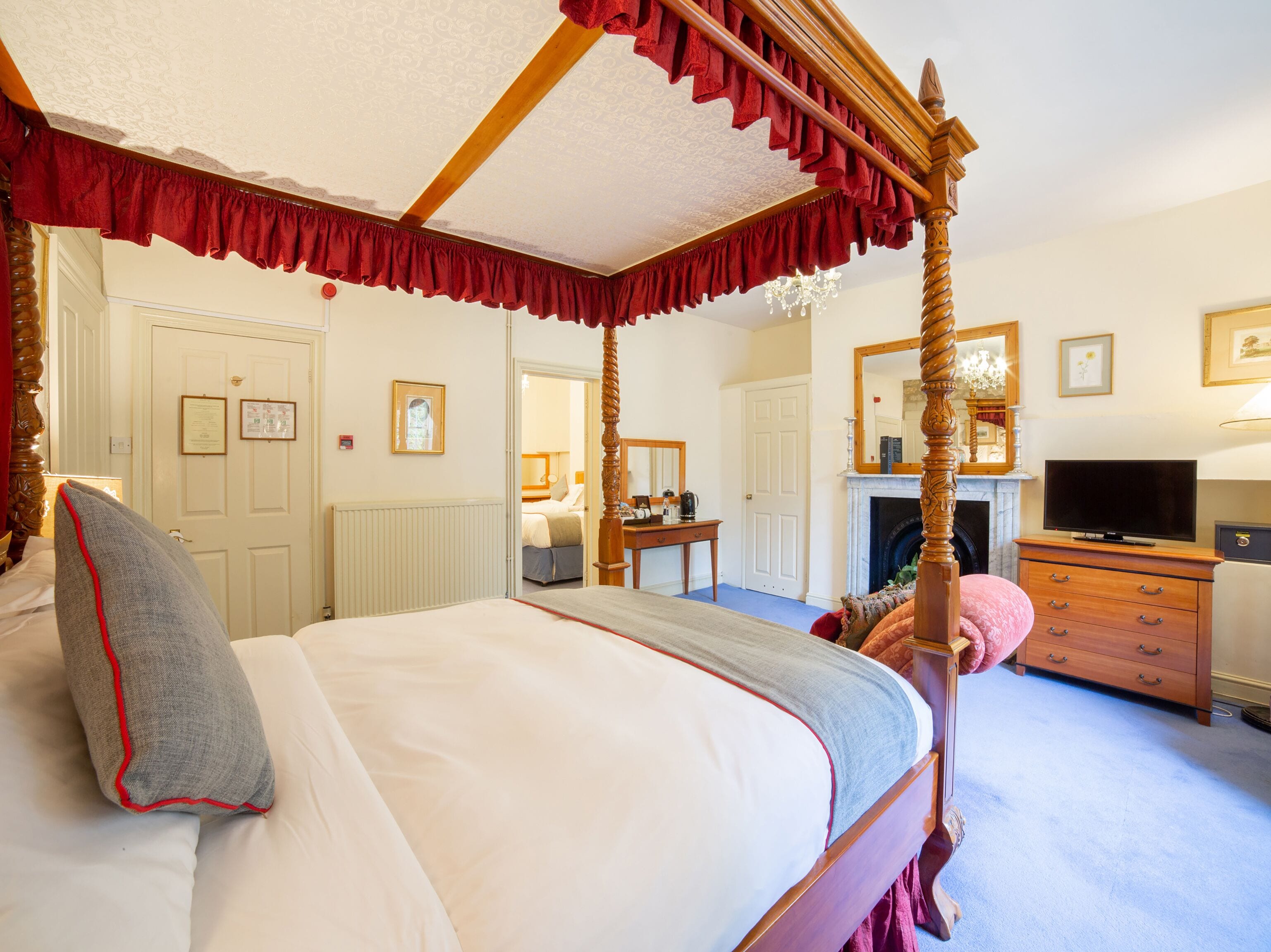 A four-poster bed with a red valance, a television on a wooden dresser, and a fireplace in the background.