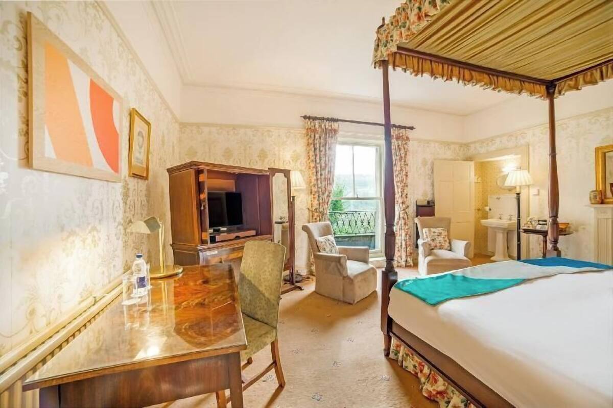 A hotel room with a four-poster bed, a desk with a chair, a television, a chair by the window, and a bathroom with a sink and mirror.