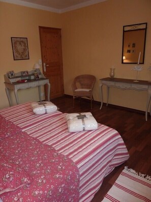 A room with a bed, a wooden desk, a chair, and a small table with a lamp.