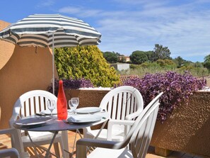 Una mesa dispuesta con sillas blancas y un paraguas a rayas, rodeada de setos cuidados y flores moradas.
