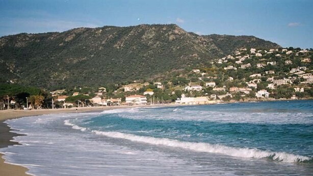 A beach with waves, a mountain in the background, and buildings on the hillside.