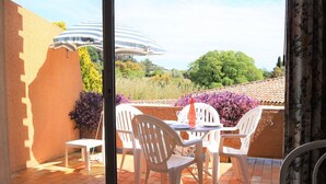 A terrace with white plastic chairs and a table, overlooking a garden with purple flowers and a building with a striped roof.