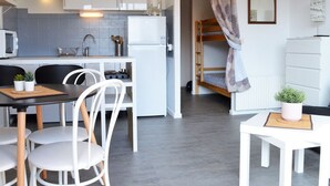 A compact kitchen with white cabinets, a microwave, and a sink. There is a dining area with a table and chairs, and a bunk bed with a patterned curtain.