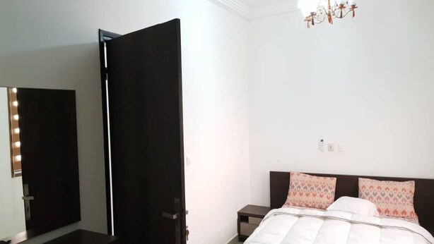 A bedroom with a bed, a nightstand, a chair, a mirror with lights, and a chandelier. - Résidence Esther 2 (Abidjan)