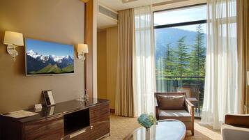 A hotel room with a large window offering a mountain view, a flat-screen TV mounted on the wall, a wooden console table, a sofa, and a glass coffee table.
