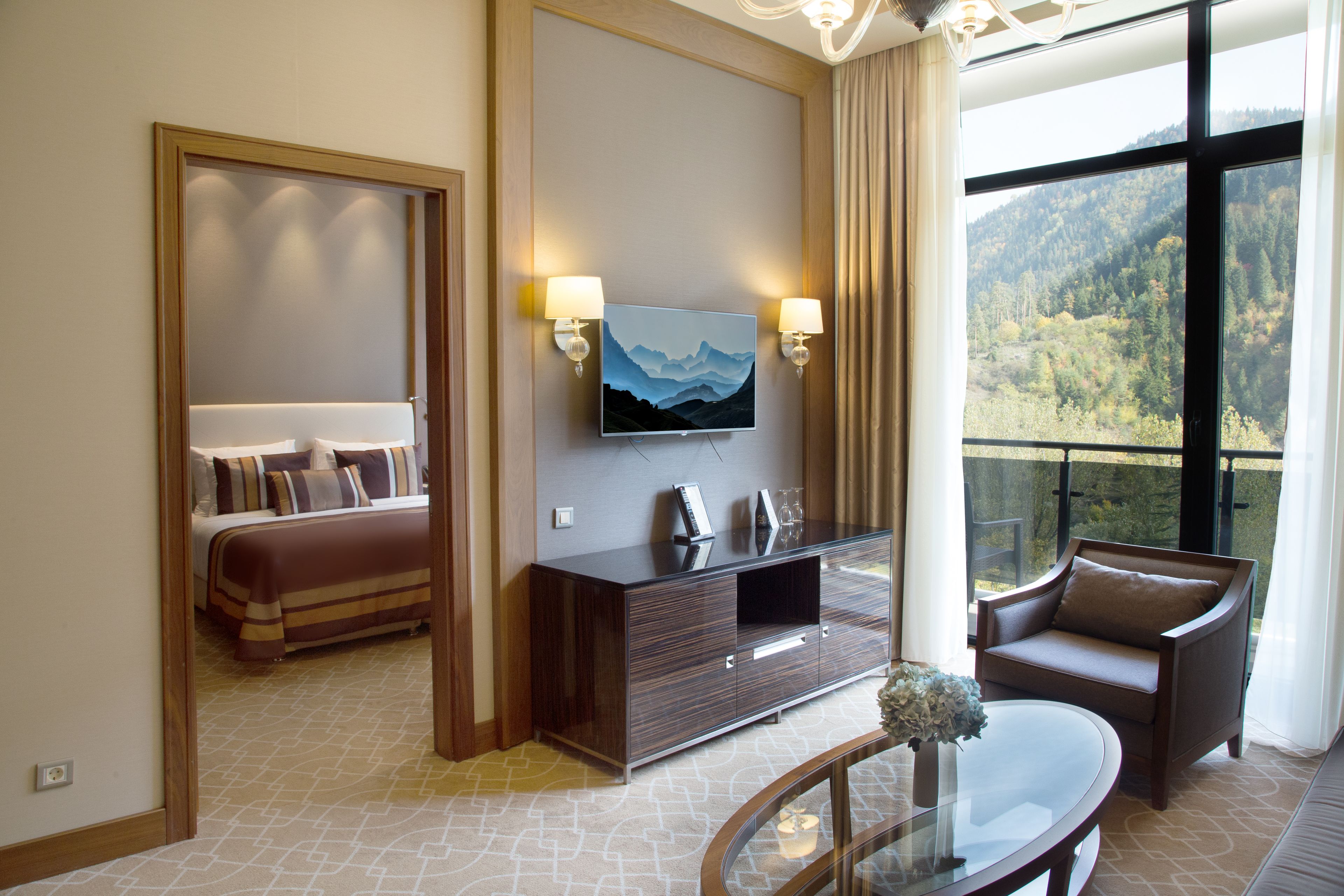 Une chambre d’hôtel avec un lit, un téléviseur posé sur une commode en bois, un canapé et un balcon offrant une vue sur la montagne.