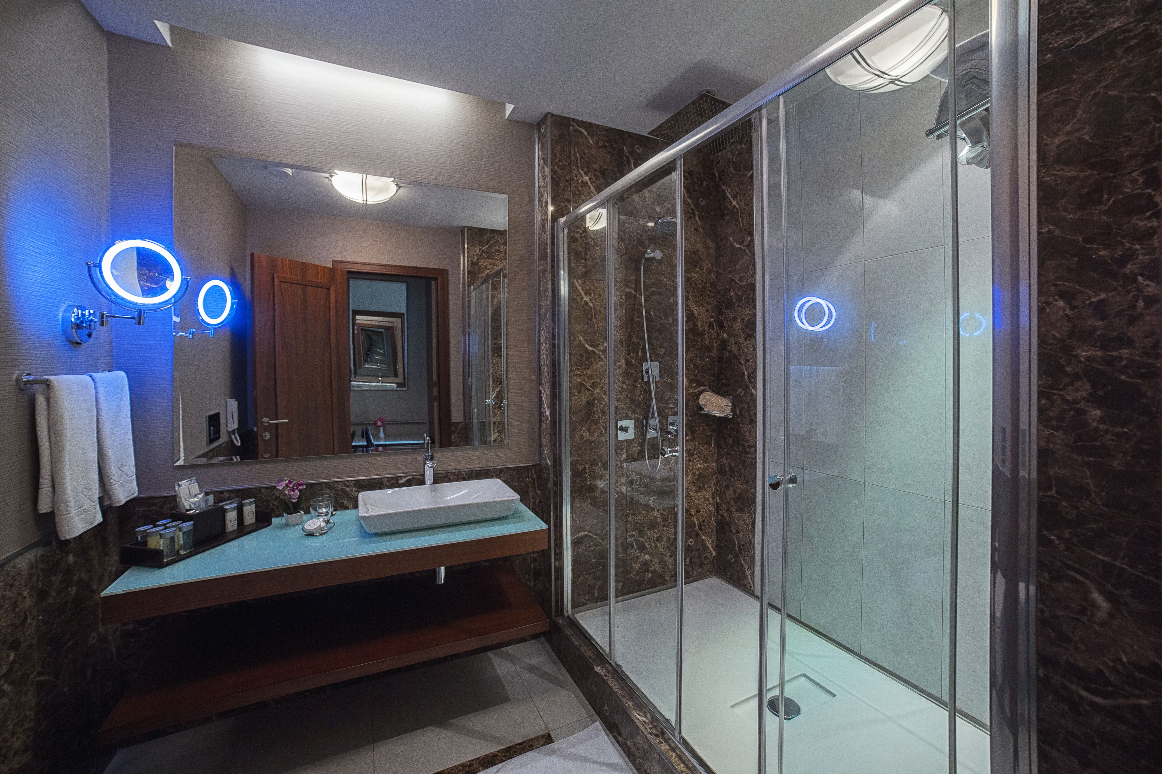 Un baño moderno con ducha de vidrio, tocador de mármol y un espejo grande con luces circulares.