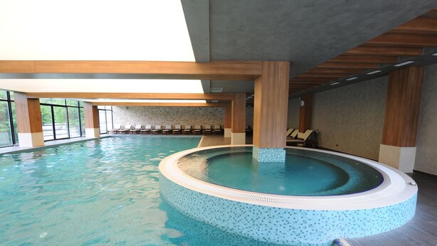 Una amplia piscina interior con un jacuzzi, vigas de madera y una pared de piedra al fondo.