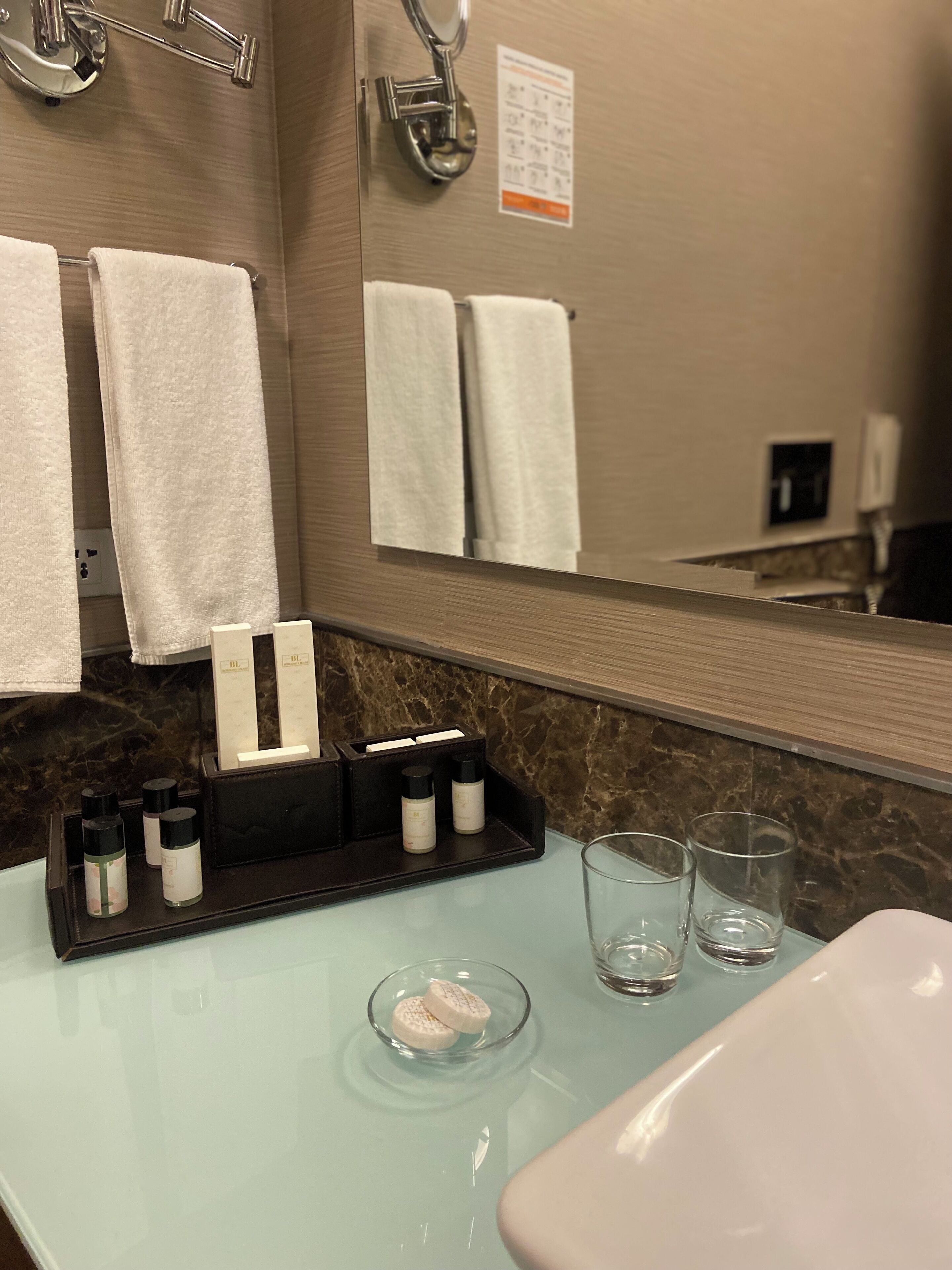 A bathroom with a marble countertop, a sink, and a mirror. There are towels hanging on the wall, a soap dispenser, and a glass of water.