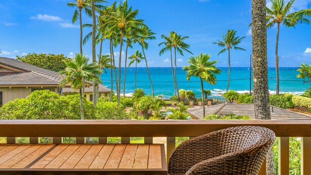 A wooden deck with a wicker chair, overlooking a coastal view with palm trees and a clear blue sky.