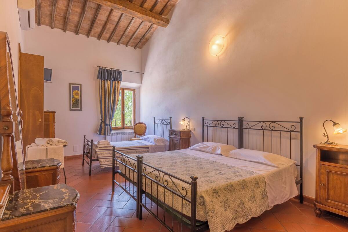 Een slaapkamer met een bed, een stoel, een houten kast, een raam met gordijnen en een plafond met zichtbare balken.