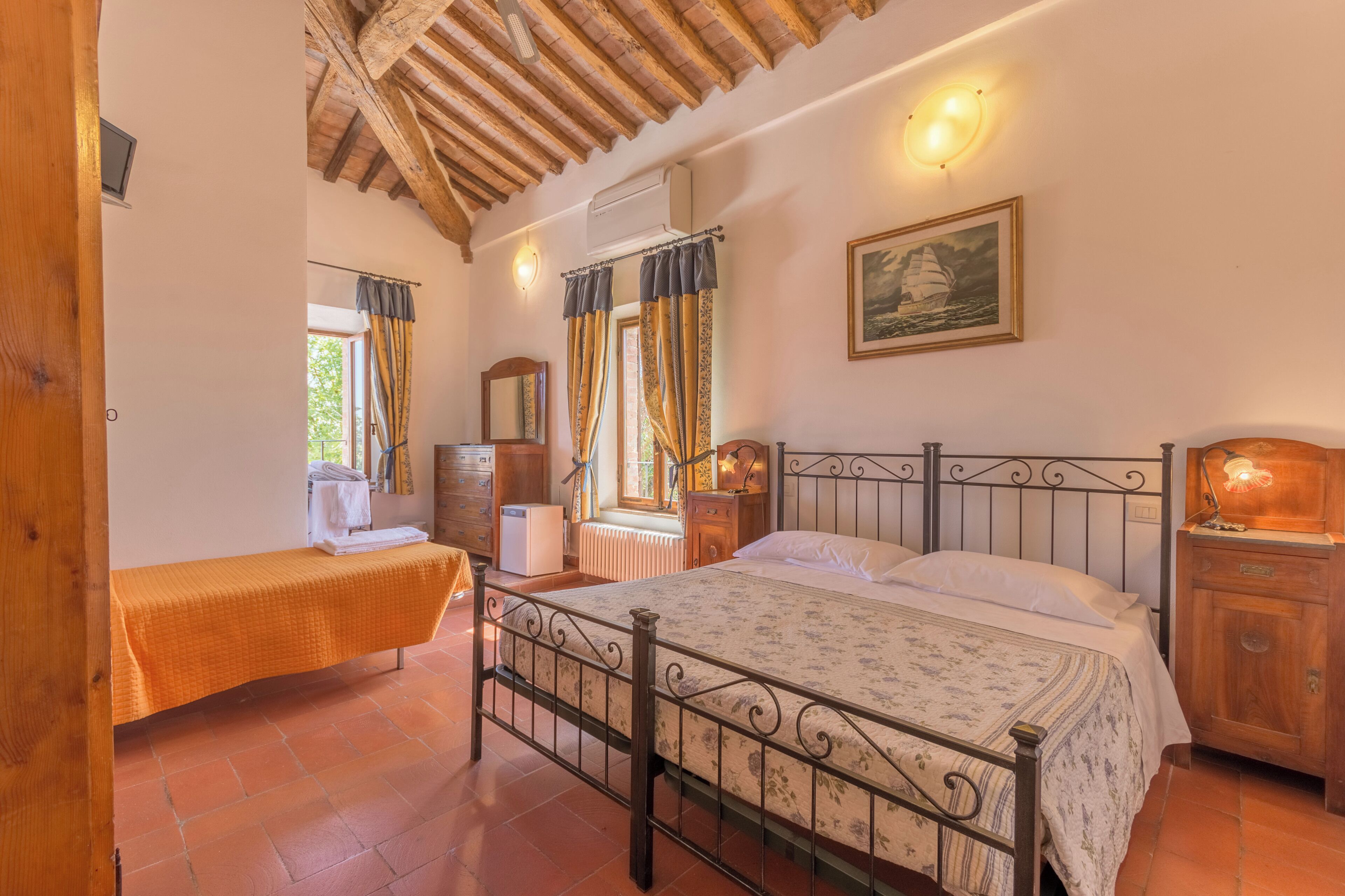 A bedroom with a bed, a nightstand, a dresser, a window with curtains, a painting on the wall, and a wooden ceiling.