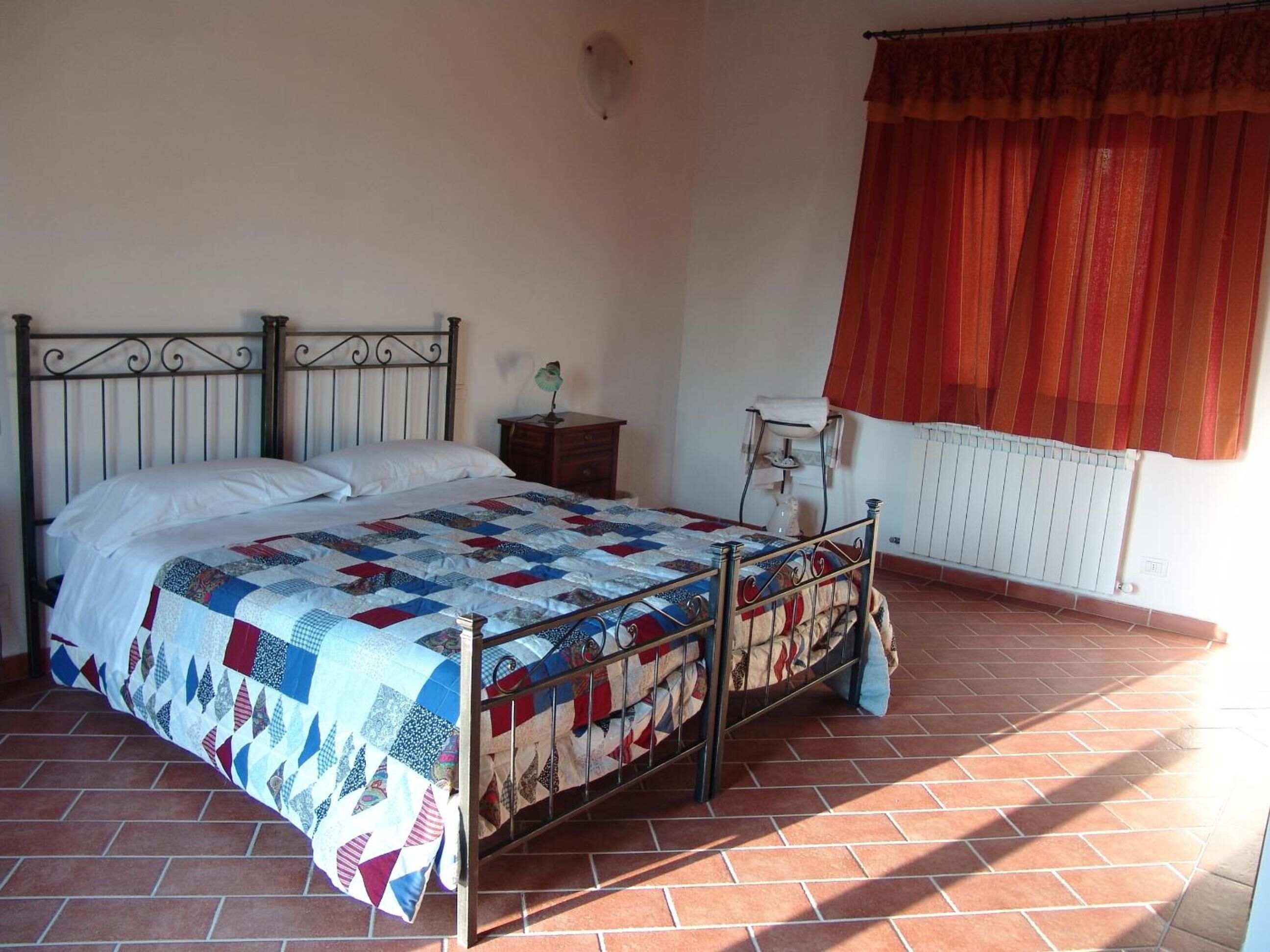 Un dormitorio con estructura de cama metálica, colcha acolchada, mesita de noche con lámpara, radiador y ventana con cortina roja.