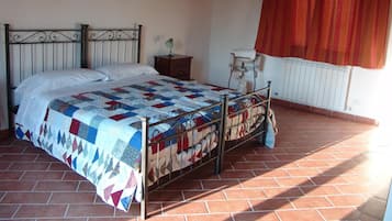 Un dormitorio con estructura de cama metálica, colcha acolchada, mesita de noche con lámpara, radiador y ventana con cortina roja.