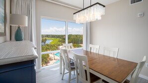 Un espace repas avec une table en bois et des chaises blanches, de grandes fenêtres donnant sur un terrain de golf et une piscine, et un lustre moderne.