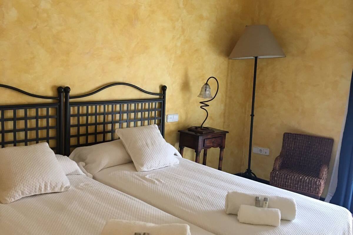 A double bed with two pillows and two towels, a lamp, a chair, and a small table in a room with yellow walls and wooden beams.