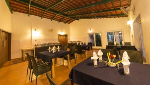 A dining area with tables set for service, a kitchen in the background, and a high ceiling with exposed beams. - Eka Resorts (Sakleshpur)