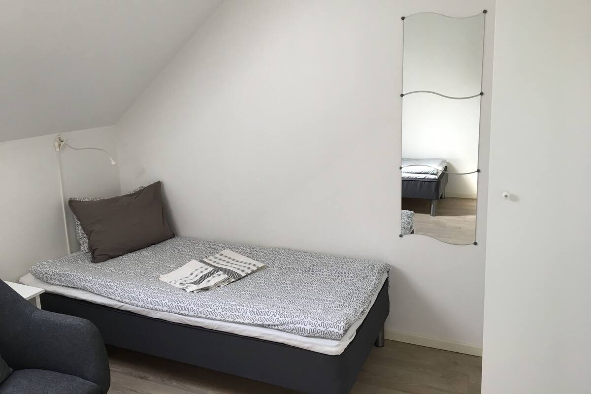 A bedroom with a bed, a chair, a bedside table, a mirror, and a door.