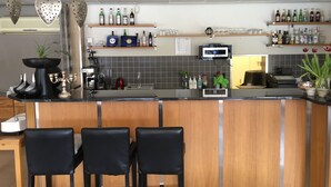 Eine Bar mit drei schwarzen Barhocker, einer Arbeitsplatte mit verschiedenen Flaschen und einer Kaffeemaschine, sowie einer herzförmigen Dekoration an der Decke.
