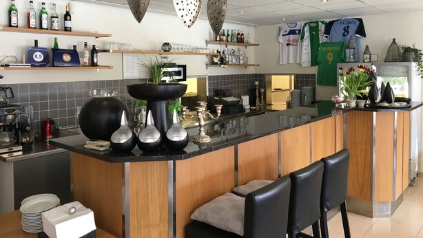 Eine moderne Bar mit Holztresen, schwarzen Barhocker und einer Auswahl an Flaschen und Gläsern auf Regalen.