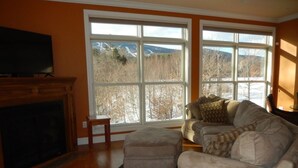 A living room with a fireplace, a sofa set, and large windows with a view of trees and mountains. - 2026-27 dates available! Mountain biking or hiking the Green Mtn, or ski Okemo! (Ludlow)