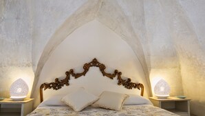 A bedroom with a vaulted ceiling, ornate headboard, two bedside lamps, and a floral-patterned bedspread. - Domus Dejana (Gallipoli)