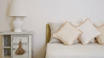 Une chambre à coucher avec un lit blanc, deux oreillers, une table de chevet blanche avec une lampe, et une petite commode en bois ornée d’une frange.