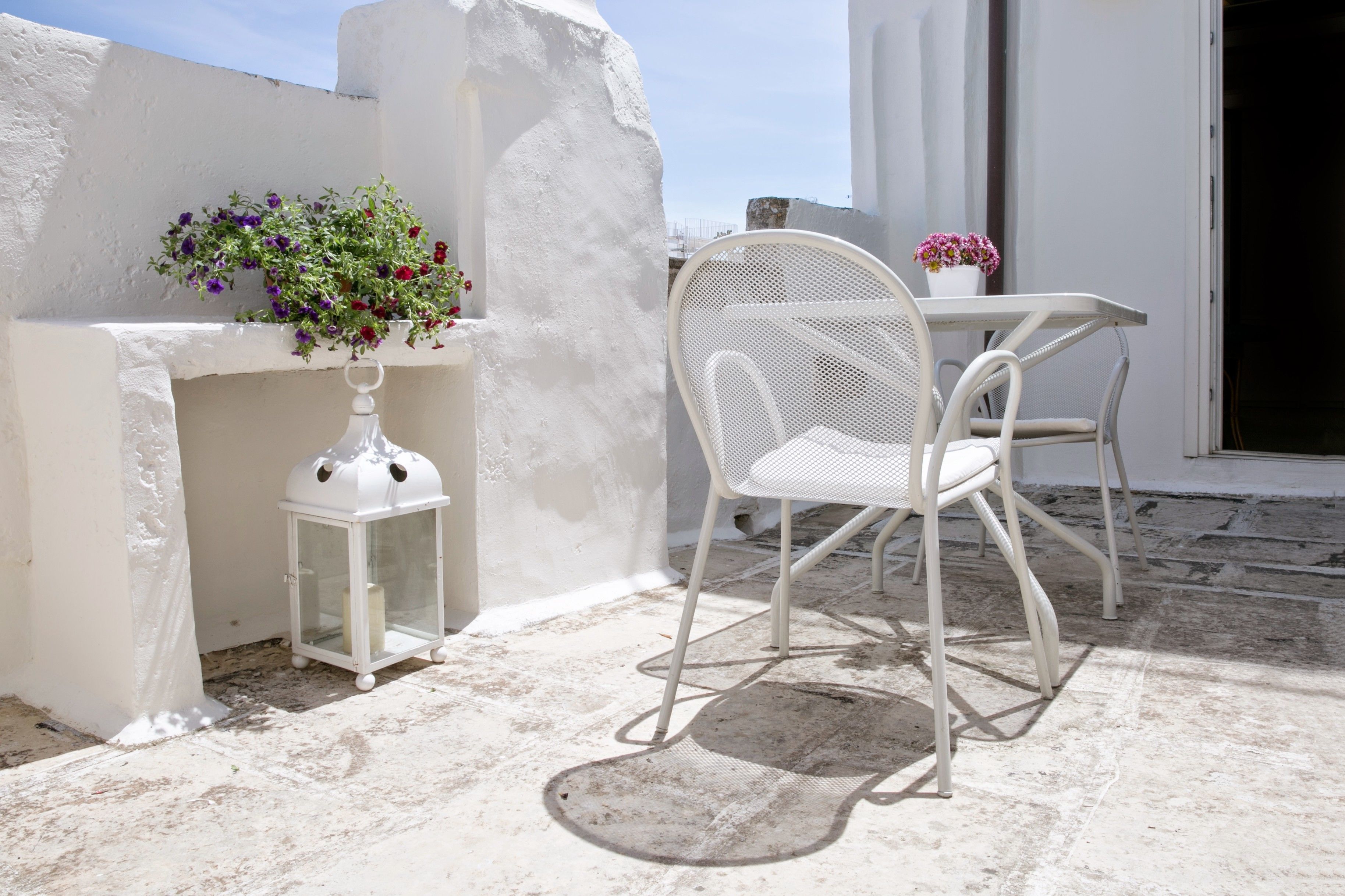 Un área de estar al aire libre con una silla y mesa blancas, un farolillo blanco pequeño y una maceta con flores.