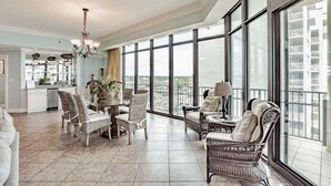 Ampla sala de estar com mesa de jantar, cadeiras de vime, sofá e varanda com vista para a cidade.