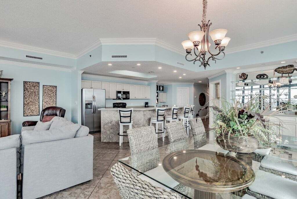 A spacious living area with a dining table, chairs, and a sofa. There is a kitchen in the background with white cabinets and a refrigerator.