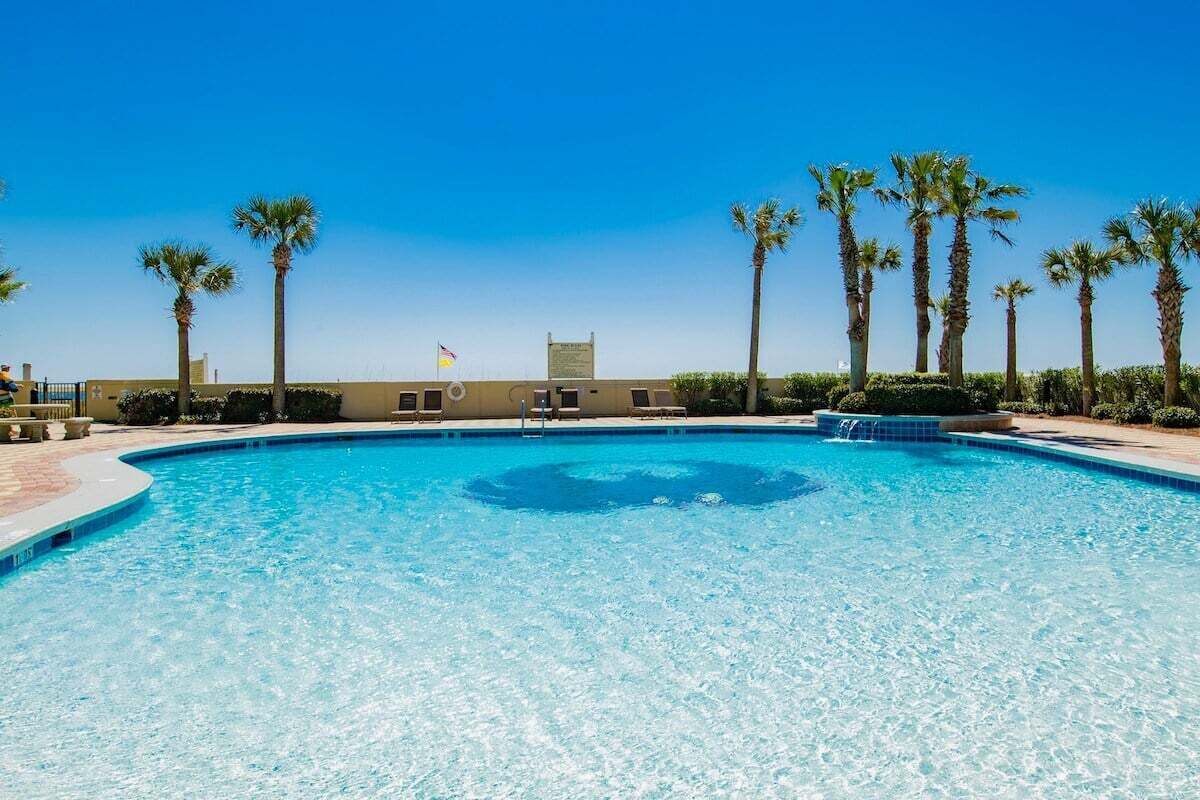 A clear blue pool surrounded by palm trees and a building with a flag.