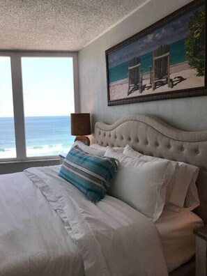 Une chambre avec un grand lit, une vue sur l’océan et un tableau représentant des chaises de plage accroché au mur.