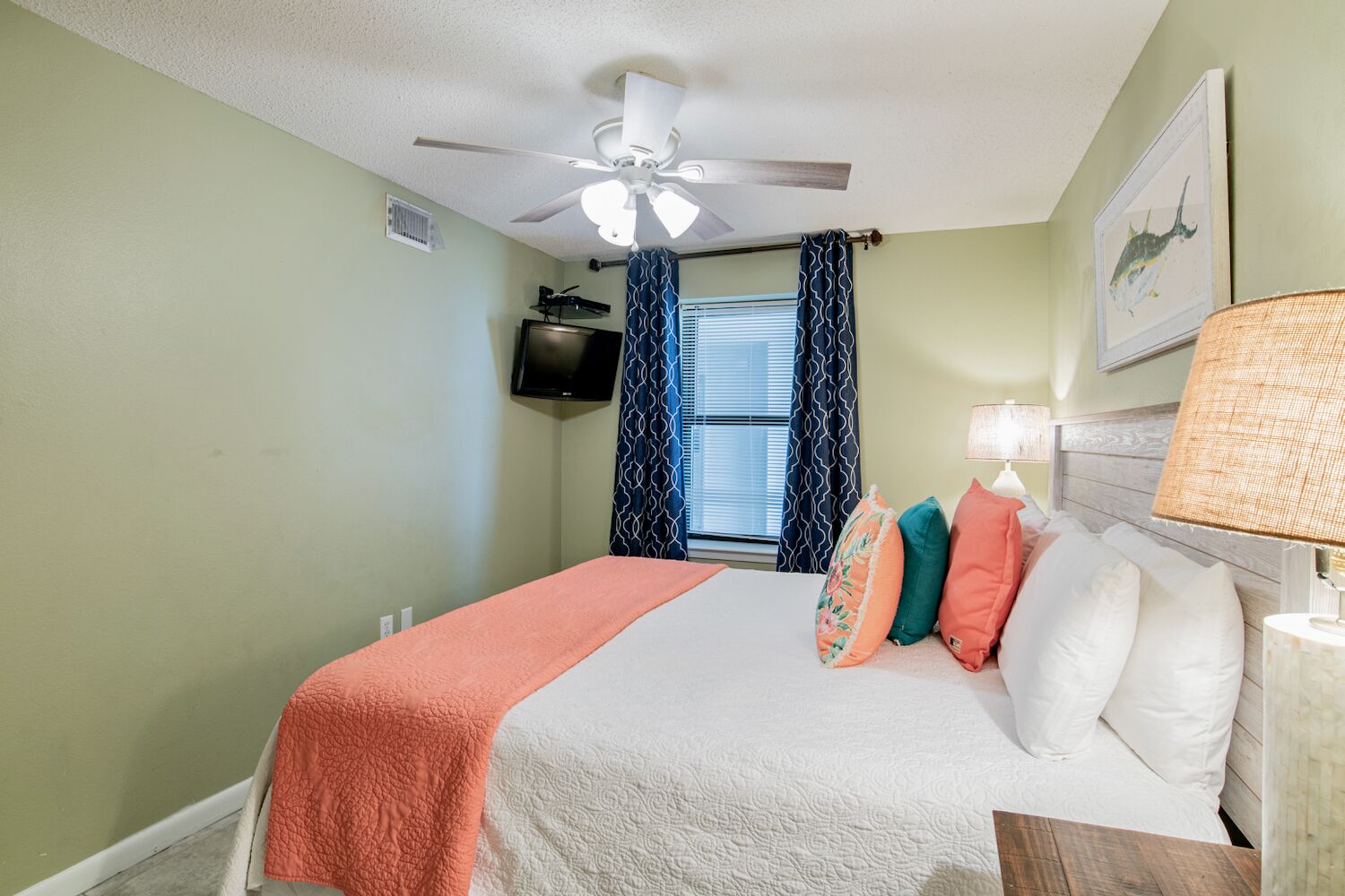 A bedroom with a bed, a ceiling fan, a TV, and a window with patterned curtains.