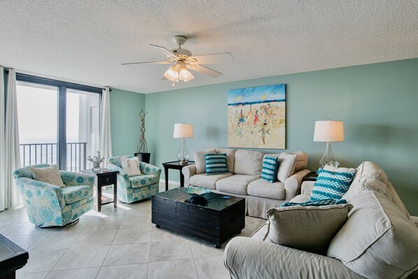 A living room with a teal wall, a ceiling fan, a large painting, and two lamps. - Phoenix VI 6903 | Updated, Clean Beach Front OBA Condo! One free covered parking included! (Orange Beach)