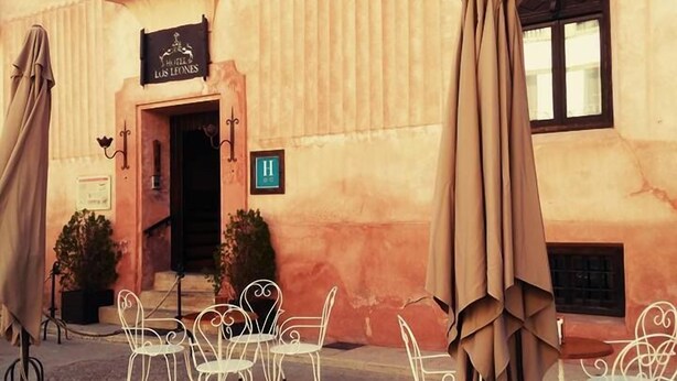 Un área de estar al aire libre con sillas de metal blanco y sombrillas, la entrada de un edificio con un letrero que dice "Hotel Los Leones" y una señal que indica una entrada accesible para personas con discapacidad.