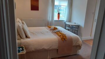 A neatly made bed with pillows and a blanket, a small table with a chair, a radiator, and a window with curtains.